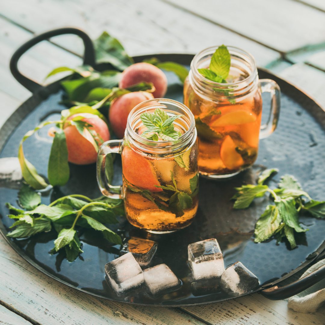 Summer refreshing cold peach ice tea with fresh mint leaves in glass jars on metal tray over rustic wooden garden table, selective focus, copy space, square crop